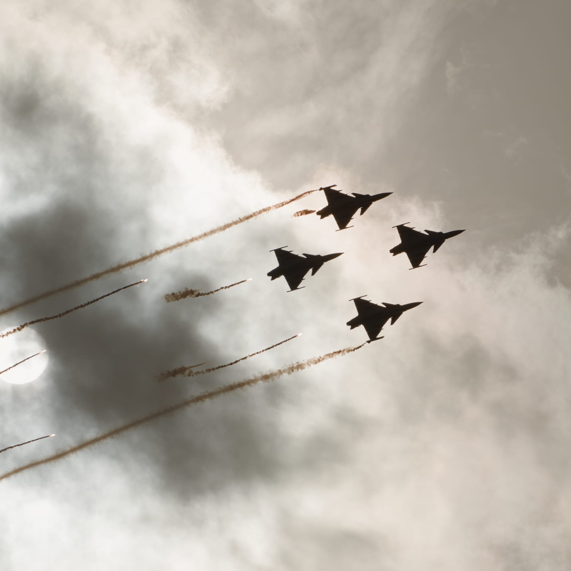 Fyra stridsflygplan i formation sett mot en molnig himmel. Alla flygplanen har fällt facklor. Bilden är tagen med väldigt hög kontrast rakt mot solen som syns till vänster i bild.
