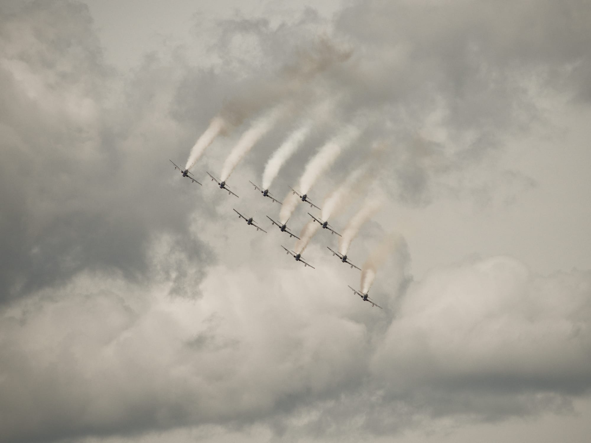 Nio flygplan som flyger i formation mot en molnig himmel. Flygplanen släpper ut vit rök.