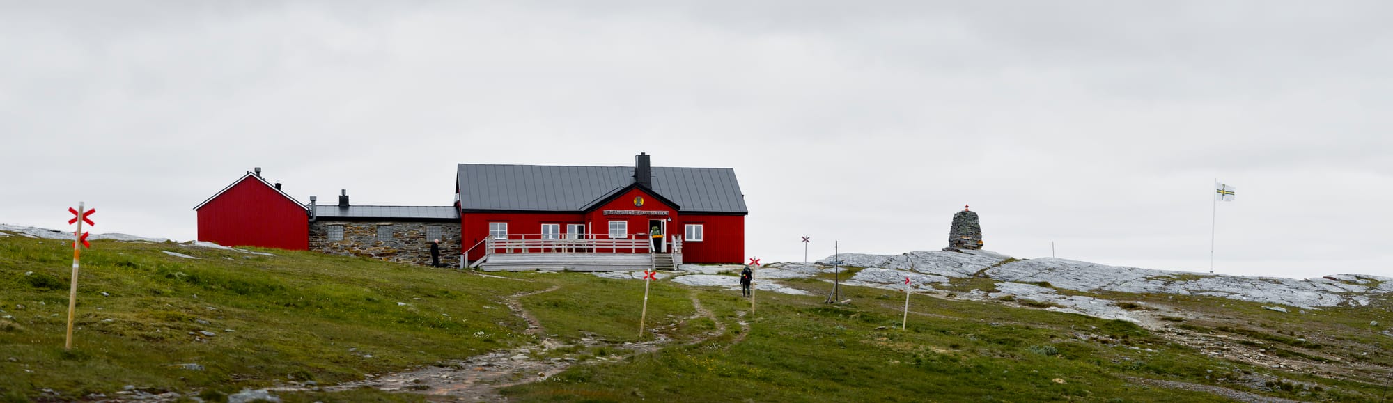 En röd byggnad på toppen av ett stenigt berg. Till höger om byggnaden syns ett stenröse och en vit, gul, blå korsflagga med STF:s logotyp i mitten